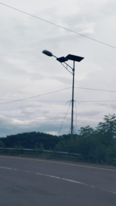 The image depicts an outdoor roadside scene in Lonbaroh, Indonesia, on an overcast day. The primary subject is a solar-powered street light, centrally positioned. It features a tall, dark pole with a single light fixture extending to the left and a rectangular solar panel mounted to the right. Several thin, dark lines, likely utility wires or cables, are visible crossing the muted, cloudy sky.
In the mid-ground, a dense stretch of dark green trees forms a natural horizon, possibly indicating a hill or forested area. A dark, paved road with white dashed lane markings occupies the foreground, running horizontally across the bottom of the frame. A silver-colored guardrail is partially visible between the road and the treeline.
The sky is uniformly grey with soft cloud formations, suggesting a cloudy or heavily overcast weather condition. No specific time of day is discernible, though it is clearly daylight. There are no people, vehicles, or explicit activities taking place, presenting a quiet, static view of the roadside environment. No visible text is present in the image.