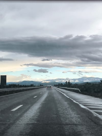 The image shows a view from the perspective of a driver on a wet highway. The road stretches forward with white lane markings visible. In the distance, a white car travels ahead in the same lane. Guardrails line both sides of the road. In the background, mountains are visible under a cloudy sky with patches of blue. The environment appears to be rural, with trees and vegetation visible along the roadside. There are no visible signs indicating this is Mursalevo, Bulgaria. There are no people visible in the frame. The focus is on the road ahead and the surrounding landscape.