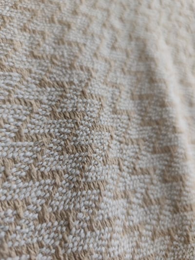 The image shows a close-up, shallow depth-of-field view of a woven fabric. The fabric has a textured pattern with alternating bands of white and beige threads. The weave appears to be a simple plain weave with some raised sections creating a subtle geometric pattern, possibly resembling small chevrons or zigzags. The lighting suggests the fabric is indoors or in a shaded outdoor area, as there are no harsh shadows. The focus is on the texture and pattern of the material, with the background blurred. There is no information about Mykulychyn, Ukraine, or any people or actions within the image.
