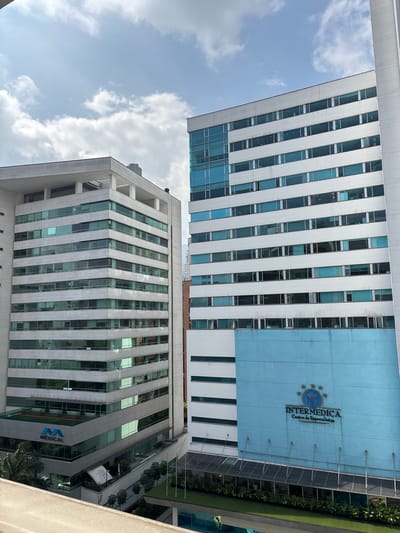 The image captures two modern high-rise buildings in Medellín, Colombia, viewed from a slightly elevated position. The building on the top is white with a blue section bearing the signage "INTERMEDICA Centro de Especialistas" and a logo with stars. The other building has the inscription "MA MEDICAL". Both structures exhibit a striped pattern created by alternating bands of windows and solid material. The sky is partly cloudy, visible in the upper-left. Greenery and part of another building is visible on the right side of the top building. The buildings appear to be located near each other, possibly within the city's urban center. There are no people or animals directly visible in the frame.