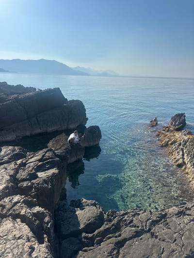 A person wearing a white shirt and dark shorts sits on a dark, rugged rock formation overlooking a body of calm, clear, blue water. The water reveals a rocky, submerged seabed. In the distance, hazy mountains rise against a pale blue sky. The scene is characterized by natural elements, specifically the rocky coastline and the serene seascape. The person is the sole human subject in the frame, positioned centrally on the rocks. The overall environment suggests a tranquil, coastal setting, likely in the vicinity of Drobnići, Montenegro, given the distinctive mountainous backdrop and clear waters often found in the region.