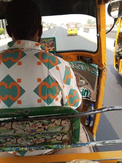 The image depicts a forward-facing perspective from inside a moving yellow three-wheeled vehicle (Keke Napep) in Gora, Nigeria. The back of a person, presumably the driver, is prominent in the foreground. This individual wears a short-sleeved, collared shirt patterned with repeating orange 'C' shapes within green diamonds and smaller orange squares on a white background. The vehicle's interior is predominantly yellow. The dashboard features a green decorative panel displaying "SAMA SAMA" alongside cartoon animal illustrations, and a strip below the windshield shows various cartoon characters. A blue-capped plastic water bottle rests in a holder on the right side of the driver's area. The passenger seat in the immediate foreground, visible from behind, is green and worn, upholstered with a fabric featuring Mickey Mouse characters and the word "Richman."
Through the windshield, a wide, multi-lane paved road with white lane markings extends into the distance under a bright sky. Another yellow three-wheeled vehicle is observed moving forward in the same lane ahead. Smaller vehicles are faintly visible further down the road. To the immediate right, a portion of another yellow three-wheeled vehicle is visible, indicating parallel movement. The roadside includes low concrete barriers and sparse vegetation in a clear, daytime environment.