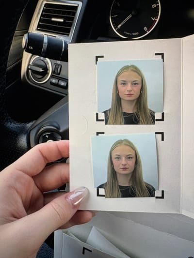 The image displays a close-up perspective of a human hand, specifically the thumb and index finger, holding a white cardstock sheet containing two identical passport-style photographs. The hand exhibits manicured nails with a clear or light pink gel polish. Each photograph features a young female with long, straight, light blonde or light brown hair, a fair complexion, and a neutral expression, facing directly forward. She is wearing a dark, possibly black, crew-neck top. The background of both photographs is a plain, light blue or grey. The cardstock has black L-shaped markings framing the corners where each photo is positioned.
The background of the scene reveals the interior of a vehicle, consistent with a Range Rover model. On the left, a section of a black steering wheel with silver trim and integrated control buttons is visible. Above the steering wheel, a rectangular air vent with horizontal slats and a circular light control dial with various settings are present. To the upper right, a portion of the vehicle's tachometer or RPM gauge, marked up to '8', is visible. The environment is an enclosed vehicle cabin, illuminated by what appears to be ambient daylight.
Regarding the location in Electoral Area A, Canada, the visual evidence within the image is insufficient to determine a precise geographical location. The scene depicts a generic vehicle interior without any specific external landmarks, distinct signage, or unique contextual elements that would allow for a forensic identification of an exact city or specific locale within Electoral Area A.