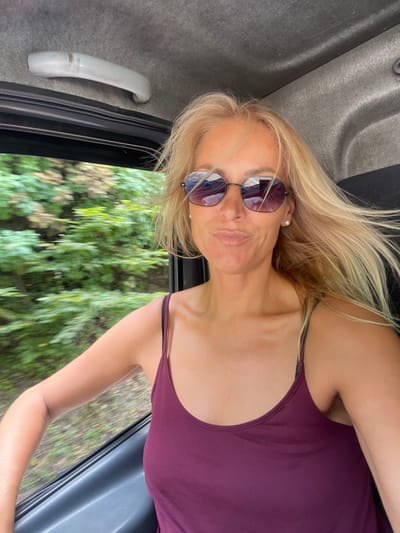 In Agoy, Russia, a fair-skinned woman with blonde hair and sunglasses is pictured from the chest up, taken from inside a vehicle. She is wearing a burgundy tank top and a pearl earring. The background shows blurred green foliage, indicating movement, and a gray interior of a vehicle with a grab handle on the ceiling. The woman is looking at the camera with her lips pursed and her head tilted slightly.