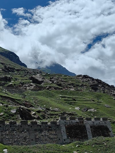 The image depicts a mountainous landscape in Yari-Khoksar, India. The foreground features a stone retaining wall with visible brickwork and concrete elements, supporting a grassy slope. The slope is covered in green grass and scattered rocks, with evidence of terracing in some areas. Further up the slope, large boulders and rocky outcrops are prominent. The background reveals more rugged mountain terrain, with some areas shrouded in clouds. The sky above is a mix of blue with scattered white, puffy clouds. There are no people or animals visible in the image.