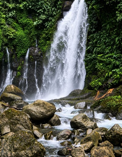 The image depicts a majestic waterfall cascading down a lush green cliff face, surrounded by dense tropical vegetation. The water plunges into a rocky riverbed filled with smooth, worn boulders and smaller stones. The scene is dynamic, with the white spray of the waterfall creating a mist that obscures some of the background details. The overall impression is one of natural beauty and untamed wilderness, with the powerful force of the water evident. The specific location in Gunung Ambat, Indonesia, suggests a tropical environment, likely characterized by high humidity and abundant plant life, which is consistent with the visual cues in the image. There are no people or animals visible in the frame, focusing solely on the natural elements.