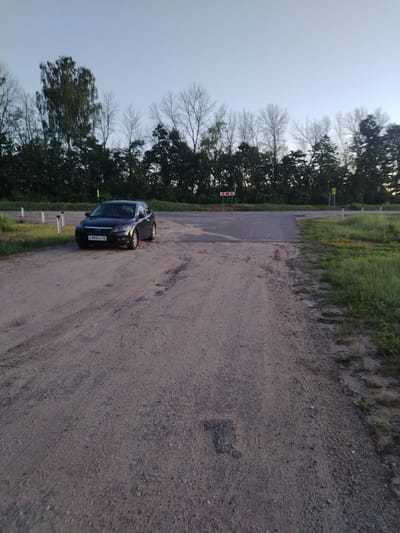 This image depicts a rural road intersection in Petrovskoye, Yaroslavl Oblast, Russia, under soft ambient light, likely dawn or dusk. The foreground is dominated by a wide, unpaved dirt and gravel road exhibiting numerous tire tracks. A dark-colored Ford Focus hatchback, likely grey or black, is parked on the left side of this dirt road, angled slightly towards the right of the frame. Its front license plate, "c 956KC 76", is clearly visible, with "76" indicating the Yaroslavl Oblast region.

The dirt road in the foreground intersects with a narrower, paved asphalt road positioned further back in the central midground. To the left of the paved road, a red and white chevron warning sign, indicating a sharp right turn, is mounted on a post. On the right side of the paved road, white cylindrical road delineators or markers are present along the grassy shoulder. A yellow pole or marker is also visible beyond the paved road, closer to the tree line.

The background consists of a dense line of trees, predominantly dark green deciduous foliage, though some trees exhibit bare branches, possibly indicating seasonal variation or different species. The sky above is a clear, light blue-grey, contributing to the soft, even illumination of the scene. No individuals or active movements are observed.