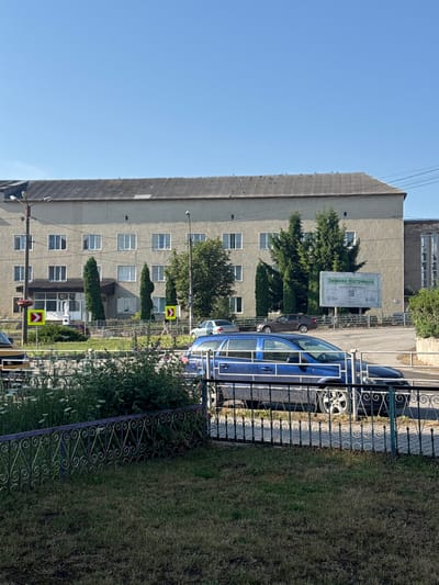 In Husiatyn, Ukraine, a blue station wagon is parked in front of a grey, multi-story building. The building has numerous windows and a grey, corrugated roof. In front of the building, there is a sidewalk, some trees, and a grassy area. Two yellow signs with red arrows are visible on poles. A large billboard is also present. The sky is clear and blue. A decorative metal fence is in the foreground, with a grassy lawn behind it.