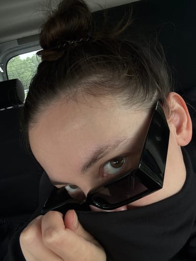 A person with fair skin and dark brown hair pulled into a bun is shown in a close-up, tilted portrait orientation. They are wearing black sunglasses. Only one eye is fully visible, and it is brown. The person is wearing a black garment. Part of their ear is visible. A hand is holding the sunglasses. A window is visible in the background with green trees. The location is not discernible from this image.