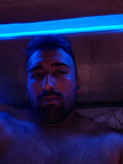 The media file shows a close-up, vertically oriented image of a person lying down and looking towards the camera. The scene is dimly lit, with prominent blue and red lighting casting strong shadows. The person appears to be of adult age, with a beard and dark hair. Their eyes are open, and their gaze is directed at the viewer. The background is dark and indistinct, but hints of a textured surface, possibly upholstery or fabric, are visible behind the person. A bright blue light source runs vertically along the right side of the frame, illuminating a portion of the scene and the person's face. The lighting creates a dramatic and moody atmosphere, with the blue light dominating the left side of the image and the red light highlighting the right side of the face. No specific activity or event is clearly depicted beyond the person lying down and looking at the camera. The overall impression is one of introspection or a posed portrait with dramatic lighting. The location context of Evere, Belgium, is provided but does not offer any discernible visual cues within the image itself. There is no visible text in the media file.
