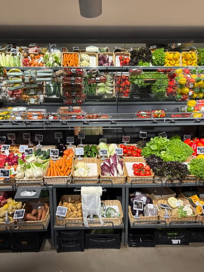 This is a vertical, eye-level, medium shot of a well-stocked produce aisle in a grocery store. The aisle is filled with a variety of fresh fruits and vegetables displayed in wicker baskets and plastic packaging on metal shelving units.
The scene depicts an abundance of colorful produce, including red and yellow bell peppers, green lettuce, radicchio, tomatoes, carrots, onions, garlic, potatoes, sweet potatoes, fennel, cauliflower, and various herbs. Some items are loose in baskets, while others are packaged in plastic. Price tags are visible on some of the displays.
The lighting suggests an indoor environment, likely a supermarket, with bright overhead lights illuminating the products. The overall impression is one of a vibrant and organized display of fresh groceries. The location is indicated as Urtijëi - St. Ulrich in Gröden - Ortisei, Italy. There is no visible activity of people in the image, but the context implies a shopping environment.
