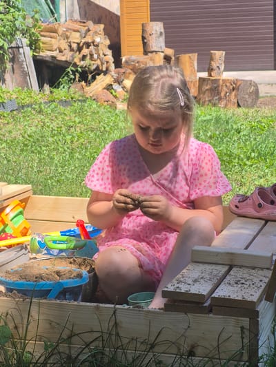 A young girl is playing with sand in an outdoor sandbox. She is wearing a pink dress with a heart pattern and a pink Croc shoe is visible on her right foot. The sandbox is made of wood and filled with sand and various plastic toys, including shovels and buckets in bright colors. The girl is holding a clump of wet sand in her hand. The background shows a grassy area, some stacked firewood, and a brown wooden fence or wall. The scene appears to be set on a sunny day, judging by the bright lighting and the shadows. The overall mood seems to be one of childhood play and enjoyment. The location is identified as Švenčionys, Lithuania. There is no visible text in the image.