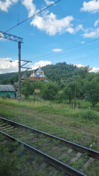 This is a daytime, eye-level, medium shot captured from a moving vehicle, likely a train, showing a pastoral scene near the village of Yasenytsia-Zamkova, Ukraine. In the foreground, parallel train tracks run diagonally from the bottom left towards the center right, with overgrown grass and vegetation growing between the wooden and concrete sleepers. Beyond the tracks, a grassy slope rises, dotted with trees and several buildings. The most prominent structure is an Orthodox church with an orange facade and a multi-domed, blue-tiled roof, topped with golden crosses. To the left of the church is a small, teal-colored building with a gray, corrugated metal roof. Further back, a dense, green forest covers a hillside, reaching up to a blue sky with scattered white clouds. Overhead, power lines and support structures are visible, indicating nearby infrastructure. The overall atmosphere is serene and rural, characteristic of the Ukrainian countryside.