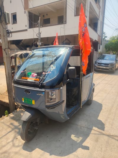 The image depicts a street scene in Anantapur, India, under direct sunlight, casting distinct shadows. A grey and black Mahindra three-wheeled commercial vehicle is centrally parked on the left side of a paved road. The vehicle displays "Mahindra" and "UDO" on its front, and its green license plate reads "AP39W N5140" with a "T" indicating transport use. Two bright orange flags, each featuring an image of Lord Rama and "जय श्री राम" (Jai Shri Ram) in Devanagari script, are prominently affixed to the vehicle's right side, extending from its roof. The windshield is adorned with several stickers, including text in Telugu script and a small green frog figure. The driver's side door is open, revealing a black seat within.

To the left and slightly behind the Mahindra vehicle, an older, yellow and black auto-rickshaw is partially visible, obscured by a utility pole heavily laden with electrical wires. Further down the street, a grey Kia Seltos SUV with license plate "AP39DC9118" is parked, facing away from the camera.

In the background, a multi-story residential building with white and light brown facades features multiple windows, balconies, and an external staircase. Utility poles line the left side of the street, one of which supports an external air conditioning unit. An open drainage ditch runs parallel to the road on the left. The paved street surface shows minor wear. No individuals are present within the frame.