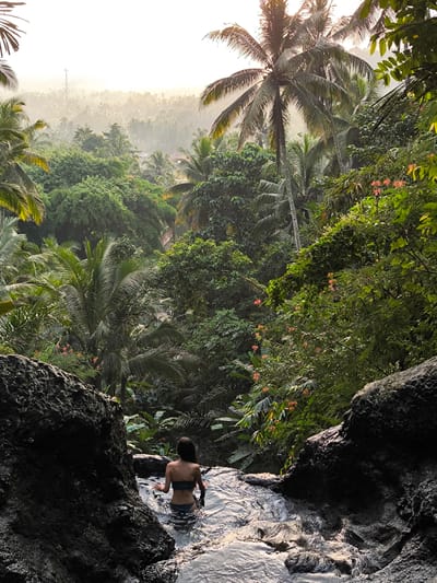 A medium-shot outdoor photo depicts a woman in swimwear standing in a shallow stream in the Sidemen, Indonesia, landscape.
The woman, viewed from behind, faces away from the viewer, standing in water that reaches her mid-thigh. She wears a bandeau-style top and matching bottoms. Her long, dark hair falls down her back.
The stream is framed by dark, jagged rocks on either side, creating a natural pool. The water appears clear and reflects the surrounding light.
The background is dominated by dense tropical foliage, including various trees and plants. Palm trees rise high above the other vegetation. In the distance, a hazy view of the landscape extends, suggesting a high elevation or a humid atmosphere. A cell tower is slightly visible in the distance on the left. The light suggests either sunrise or sunset.