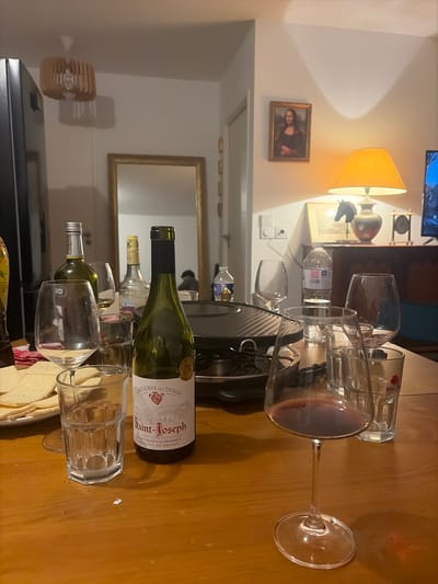 A table is set with several wine glasses, a wine bottle labeled "Saint-Joseph," and a small electrical appliance. Behind the table is a hallway with a mirror reflecting a person. A lamp with a yellow shade illuminates the scene, and a picture hangs on the wall. The room appears to be a living area, possibly a dining or living room in an apartment or house.
