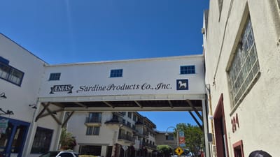 The image shows a sky bridge connecting two buildings in Monterey, California, USA. The bridge is painted white and has the words "ÆNEAS Sardine Products Co., Inc." painted on its side, along with a silhouette of a horse. Blue-framed windows are visible on the bridge and the buildings. Below the bridge, there is a street with a modern building, trees, and signs. One sign reads "MONTEREY P". A pedestrian crossing sign is visible, indicating a street. The sky is clear and blue. The overall scene depicts an urban environment with historical architecture.