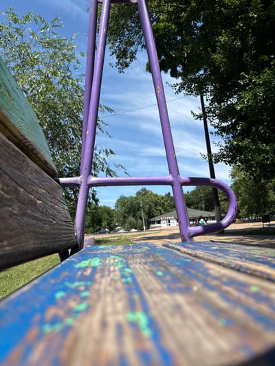 An outdoor shot captures a close-up perspective of a weathered, blue and green painted wooden bench. The bench is positioned low to the ground, with the front edge in sharp focus, showing peeling paint and wood grain. Behind the bench, a purple metal swing set frame is visible, its tubes showing signs of wear and tear with flecks of yellow paint. In the background, a paved area leads to a building with a green roof, a sign displaying Cyrillic text that reads "Я КЕБАБ" (Ya Kebab), indicating a food establishment. Several trees with lush green foliage surround the area, and the sky is clear blue with scattered wispy clouds. A dark-colored car is parked in the distance. Two indistinct figures are walking in the background near the building. The scene appears to be set in a public park or recreational area within the city of Piatykhatky, Ukraine, under bright daylight conditions.