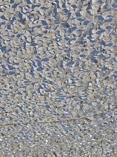 This image shows a close-up, low-angle view of a camouflage netting stretched across an outdoor area, likely a patio or seating area, under a bright blue sky. The netting is made of a lightweight material with irregular cutouts, creating a dappled effect as sunlight filters through. The pattern of the netting is a mix of light beige and grey shapes against the blue sky. The netting is supported by a grid of thin, dark cords, forming a repeating square pattern. The perspective suggests the viewer is looking upwards at the canopy. There are no people or other discernible objects visible in the frame, creating an abstract and textural composition focused on the patterns of light and shadow. The location is not specified in the image, but a general description of the visual content is provided.