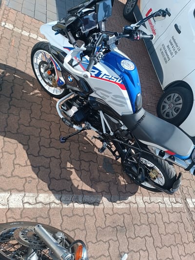 The image shows a white and blue BMW R1250GS motorcycle parked on a reddish-brown interlocking paver surface, characteristic of a public parking area or sidewalk in Germersheim, Germany. A white van with German text and a phone number is partially visible behind the motorcycle. The van's text includes "Handwerker 2.0 GmbH & Co. KG," "Werkstraße 3," "67354 Römerberg," and "Tel. 06232 68580." The motorcycle is positioned at an angle, with its front wheel to the lower left of the frame and its rear wheel towards the upper right. The scene is bathed in sunlight, casting distinct shadows from the motorcycle and a person's legs and feet at the bottom of the image. The motorcycle appears to be stationary.