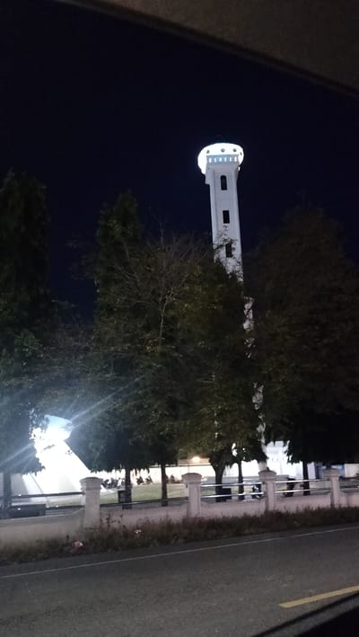 The image captures a tall, slender white tower, likely a minaret, brightly illuminated against a dark, clear night sky. The tower features small, rectangular openings and is crowned by a circular, brightly lit upper section with multiple points of light. The photograph appears to be taken from inside a vehicle, with parts of the car's interior visible at the top and bottom edges.
In the midground, several trees with varying foliage density stand between the viewer and the tower, partially obscuring it and other background structures. Below the trees, a white fence with decorative pillars runs horizontally across the frame, separating the road from a grassy area.
On the grassy area behind the fence, several motorbikes are parked, suggesting human activity or presence, possibly related to a mosque complex. Behind the trees and fence, additional brightly lit buildings are visible, consistent with a religious or public complex. A dark asphalt road with a faint yellow line is visible in the foreground. The time of day is clearly night, as indicated by the dark sky and the artificial illumination of the structures. The location is Lambarotunong, Indonesia. No discernible text is visible in the image.