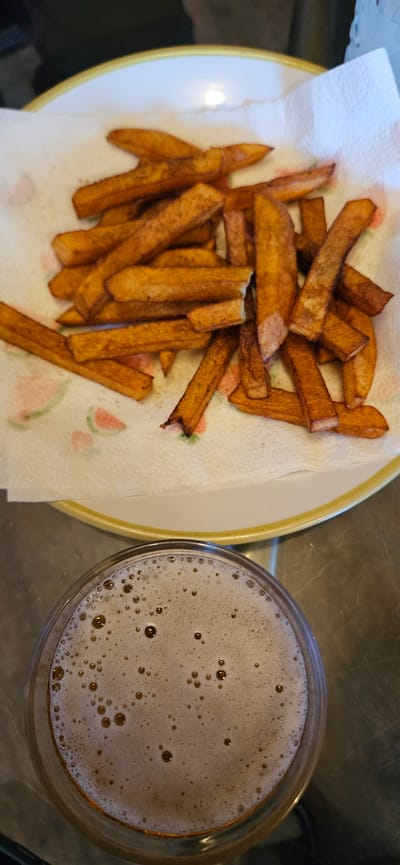 The image presents an overhead shot of a casual meal or snack, featuring a plate of chips and a drink, set on a dark, reflective surface.
On the left, a white plate with a prominent yellow rim holds a generous portion of golden-brown, thick-cut chips (fries). These chips are laid out on a white paper towel, which has faint, pastel-colored designs resembling watermelon slices in pink and green. The chips appear freshly cooked, with a crispy texture and varying shades of brown, indicating they are well-fried. They are likely placed on the paper towel to absorb excess oil.
To the right of the plate, partially cropped, is a clear glass containing a dark beverage. The drink is topped with a substantial layer of light-brown foam, dense with numerous small bubbles, typical of a beer such as an ale or stout, or potentially a dark carbonated soft drink.
The background surface appears to be a dark, smooth countertop or table, possibly metallic or stone, reflecting some ambient light. The lighting suggests an indoor setting, possibly evening. No people are visible, and there is no discernible text within the image. The scene depicts a simple, comforting food and drink pairing, common in a relaxed, home-like environment in Norwich, United Kingdom.