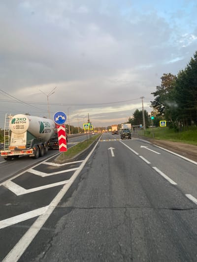 The image captures a road scene in Шароны (ангарский), Russia, during what appears to be late afternoon or early evening, with an overcast sky. The perspective is from a vehicle moving on a multi-lane road.

Dominating the left side of the frame is a large white cement truck with "ЭКО БЕТОН" (Eco Beton) printed on its side in green and black lettering, along with phone numbers. The truck has multiple axles and is positioned in one of the lanes. Adjacent to the truck, a blue circular road sign indicates a mandatory lane change to the left or right. A red and white chevron sign is also present on the right side of the truck, likely indicating a hazard or lane obstruction.

The road itself is asphalt, with clear white lane markings. In the foreground, chevron-shaped markings guide traffic. Further down the road, to the right, a pedestrian crossing is marked by yellow lines, and a yellow square sign with a pedestrian symbol is visible. A traffic light above the pedestrian crossing is illuminated green.

Several other vehicles are visible in the distance, including other trucks and cars, suggesting moderate traffic. The right side of the road is lined with trees and vegetation, with a forest visible in the background. Power lines and streetlights are also present along the roadside. The overall impression is of a highway or a major road leading into or out of a populated area. There are no people clearly visible or identifiable in the image.