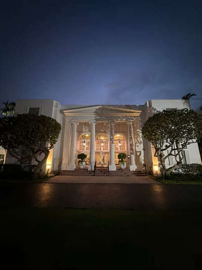 The image depicts the entrance to a white, two-story building with a grand portico supported by classical columns. The building is illuminated at night, casting dramatic shadows from its architectural features and surrounding trees onto its facade. In front of the building, there is a paved walkway leading to a set of stairs. Flanking the walkway are mature, sculpted trees. The sky above is a deep, dusky blue, indicating twilight or nighttime. The scene is located in Bal Harbour Village, United States of America.