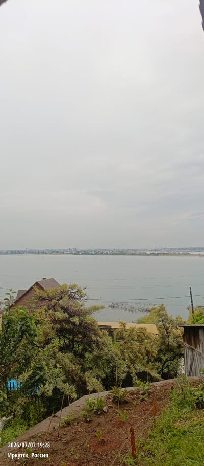 The image shows a wide view of a calm body of water, likely a river or a lake, under a hazy, overcast sky. In the foreground, there is a small wooden house with a dark roof, partially obscured by lush green trees and vegetation. The house appears to be situated on a slightly elevated slope. In the middle ground, the water stretches out towards a distant, low-lying shoreline where a city skyline with buildings and some infrastructure, possibly bridges, is visible through a light mist. The water is dotted with reeds or submerged vegetation. The foreground also includes a patch of cultivated soil with young plants and some garden stakes. The timestamp and location "2026/07/07 19:28 Иркутск, Россия" are overlaid on the bottom left of the image. There are no people visible in the scene. The overall impression is one of a quiet, somewhat diffused natural landscape with signs of human habitation.