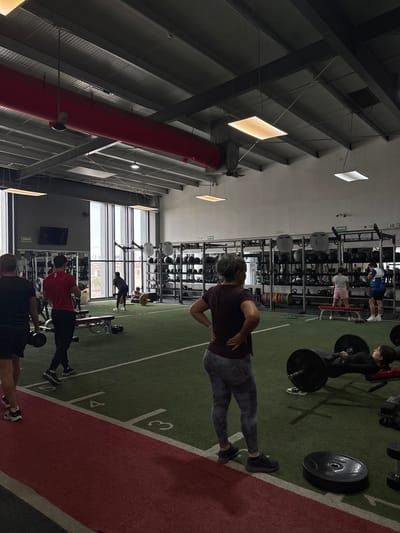 The image shows the interior of a gym in San Pedro Cholula, Mexico. Several people are exercising. At the center of the frame, a person wearing a maroon shirt and patterned leggings stands with their hands on their lower back. To the right, a person in all black appears to be lifting weights. In the background, others are using weight racks and benches. The gym has high ceilings with exposed metal beams and industrial lighting fixtures. A large window is visible on one side of the gym. The floor is a combination of green and red turf with white markings resembling a sports field. Weight plates, dumbbells, and various gym equipment are scattered throughout the space.