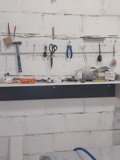 This is a top-down, full-frame shot of a collection of tools organized on a white pegboard mounted on a textured, white-painted brick wall. The pegboard extends vertically along the right side of the frame.

The tools displayed include:
- A hammer with a green and black handle.
- A screwdriver with an orange handle.
- A metal file.
- A small cloth bag.
- A pair of pliers with blue handles.
- A pair of scissors.
- A set of keys.
- A wrench.
- Several screws and nails.
- A spool of orange string with a retractable mechanism.
- Boxes of staples.
- A yellow object, possibly a ruler or level.

To the left of the pegboard, on the brick wall, there are faint pencil markings that appear to be sketches or notes. The wall itself is rough and shows the texture of painted bricks. To the far right of the frame, a curved blue pipe is visible.

The lighting suggests an indoor setting, possibly a workshop or garage, with overhead lighting casting minimal shadows. There are no people visible in the image. The overall impression is one of organized functionality, with tools neatly arranged for easy access. Some visible text includes "TOTAL," "STAPLES," "Chipboard Screw," "M4.0x30," "1000," and "D1/08." The scene is static and does not depict any ongoing activity.