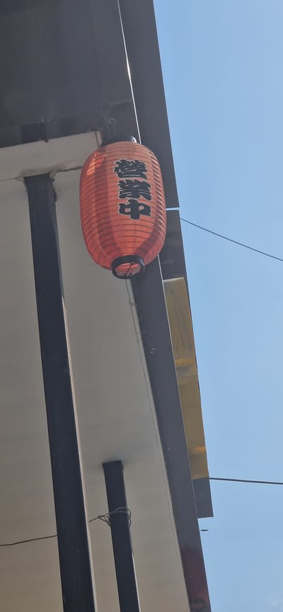 The image shows an orange Japanese lantern with black lettering hanging from the corner of a building against a clear blue sky. The lantern is cylindrical and appears to be made of paper. The lettering on the lantern reads "営業中", which translates to "Open" in English, indicating that a business is currently operating. The building has a white soffit and dark brown trim, with black metal supports visible. The context of Miass, Russia, is not directly observable from the image itself.