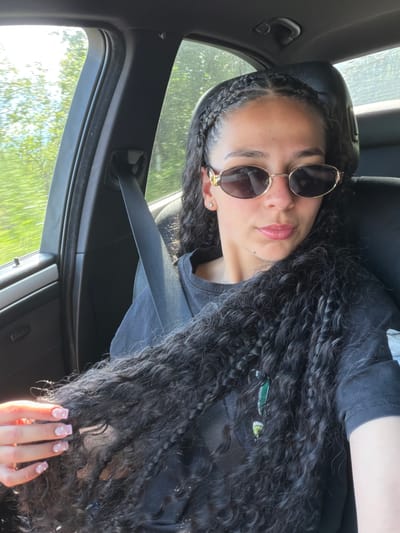 A young woman with long, dark, curly hair and braids is pictured inside a car. She is wearing sunglasses with gold frames and a black t-shirt. Her nails are painted a light pink with white designs. The woman is looking up and to her right, with a slight smile. Outside the car, trees and greenery are visible through the windows, suggesting a sunny day. The interior of the car, with a black seatbelt and dark upholstery, is also visible. The image appears to be a selfie taken from the perspective of the passenger seat.