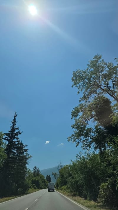 A white van drives down a paved road flanked by dense green trees under a clear blue sky with the sun shining brightly in the upper left corner. The road has faint white lines marking the lanes and appears to be in a rural or mountainous area, with a hint of a mountain range visible in the distance. The overall scene suggests a journey through a natural landscape in daylight. No specific location in Mezdreya, Bulgaria can be determined from the image alone.