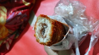 The image shows a close-up of a fried snack being held by a person's hand. The snack appears to be a breaded cylinder, possibly filled with meat or vegetables, and it is partially wrapped in clear plastic. The background is a blurry red surface, possibly a tablecloth or clothing. The image was taken in Palohlada, Indonesia.