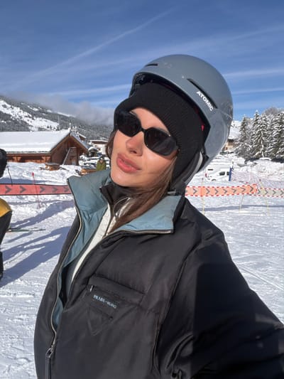 An image captured in Les Allues, France, depicts a snowy mountainous landscape under a clear blue sky. In the immediate foreground, a female individual is positioned, facing slightly left and looking towards the camera. She wears a grey Atomic ski helmet over a black knit beanie, and rectangular black sunglasses. Her attire includes a black puffer jacket with a visible light blue/grey inner collar, partially unzipped to reveal a white garment beneath and a black turtleneck. The left chest pocket of the jacket bears the "PRADA - MILANO" inscription and the Prada triangle logo. Her lips appear full and glossy.
The ground is covered in white snow, displaying distinct shadows. To the left of the primary subject, a portion of another individual in yellow ski attire and a red and white safety barrier are visible. The mid-ground features several traditional wooden chalets with snow-covered roofs, snow-laden picnic tables, and additional safety netting. Further back, a white van and other vehicles are present. The background shows dense snow-covered pine trees and majestic snow-capped mountains beneath a bright, sunny sky with sparse, high-altitude clouds.