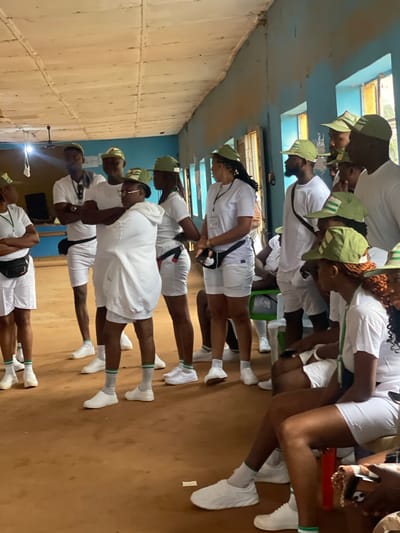 In Orun Ekiti, Nigeria, a group of young adults, likely participants in a youth service program, are gathered indoors. The room appears to be a temporary or rustic structure with a wooden ceiling and blue painted walls. The floor is a dirt or compacted earth surface. The individuals, predominantly dressed in white t-shirts, white shorts, green and white striped caps, and white sneakers, are arranged in clusters. Some are standing, while others are seated on a green bench and what appear to be white plastic chairs. Several individuals are looking towards the left of the frame, suggesting a focal point of attention or instruction. The atmosphere seems to be one of assembly or briefing, with the uniform attire implying a collective purpose. The overall scene is informal and functional, indicative of a community or service-oriented gathering in a local setting.