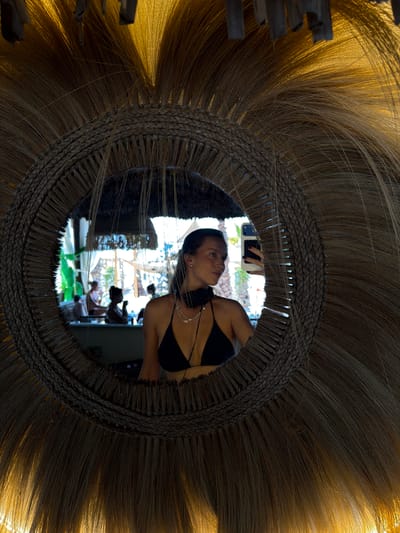 In the city of Drobnići, Montenegro, a young woman with dark hair, wearing a black bikini top and a necklace, is reflected in a large, round mirror. The mirror is framed with intricately woven straw and features a sunburst-like pattern of straw strands radiating outwards. The woman is holding a smartphone in her right hand, taking a selfie. She is wearing black headphones around her neck. In the reflection of the mirror, other people can be seen in the background, seated at tables in what appears to be an outdoor seating area with a canopy. The lighting suggests it is either sunset or sunrise, with a warm, golden glow illuminating the scene.