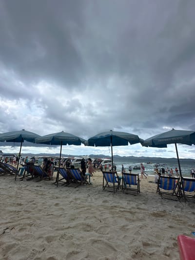 The image depicts a crowded beach in Tambon Phru Nai, Thailand, under a dark, overcast sky with heavy clouds. In the foreground, several blue beach umbrellas with "JETSKI CLUB" branding are set up on the sand, casting shadows. Rows of empty beach chairs, some with blue and striped cushions, are positioned in front of the umbrellas. Beyond the chairs, a bustling scene unfolds along the shoreline. Numerous people are visible, some walking, some standing, and some appearing to be engaged in activities near the water. A fleet of boats, including speedboats and possibly jet skis, are docked or moving in the water. The sea stretches out towards a distant, hazy coastline with hills. The sandy beach is textured with footprints and some scattered debris. The overall atmosphere suggests a busy tourist destination, though the impending weather might be deterring some beachgoers or creating a sense of urgency.