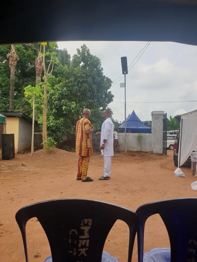 The image depicts two men standing on a dirt ground. One man is wearing a white outfit while the other is wearing a patterned brown, orange, and yellow robe. They are standing next to a building on the left side. There are a few trees around them. A canopy is visible behind the man in white. The scene appears to be in an outdoor setting, and the location is Nkwerre, Nigeria. In the foreground, there are chairs with the name "EMEK" written on it.