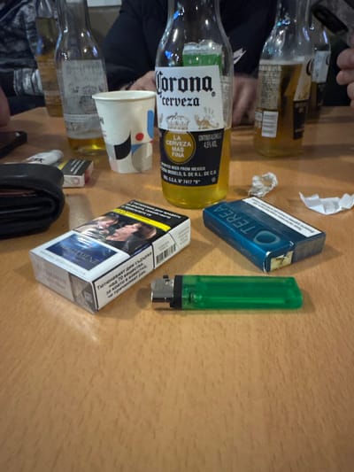On a light brown wooden surface, several items are scattered. Starting from the left, there are multiple empty glass Corona beer bottles and a paper cup with a white background and colored geometric shapes. Next to the cup is a pack of Parliament cigarettes, a pack of Terea tobacco sticks, and a green lighter. A black wallet also sits on the surface. Part of a person wearing a black shirt is visible. The environment suggests a casual gathering or setting where these items might be commonly found. Location cannot be confirmed based on the available image content.