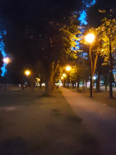 The image depicts a park scene at dusk or night, illuminated by streetlights. A paved pathway runs through the park, bordered by grass and mature trees. Several lampposts are visible, casting a warm, yellow glow on the surroundings. In the background, silhouetted figures of people can be seen walking or standing, suggesting it is a public space. One or two park benches are present along the pathway. The sky is dark blue, with hints of lighter blue peeking through the tree canopy. The overall atmosphere is calm and serene. The location is identified as a park in Stryków, Poland.