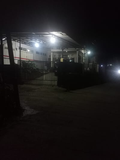 The image depicts a nighttime exterior scene in Kecamatan Pangalengan, Indonesia, focusing on a building illuminated by artificial lights. The time of day is clearly night, with the surrounding environment plunged into darkness, contrasting with the brightly lit area beneath a large, corrugated metal roof structure.
The main subject is a building complex situated behind a dark metal gate with vertical bars. The building on the left features a long, light-colored wall with horizontal red stripes or markings and multiple windows or vents, suggesting it could be a warehouse, workshop, or industrial facility. On the right, another section of the building has visible pillars, possibly indicating a porch or an office area. The entire structure is covered by an extensive metal roof, under which several bright lights are mounted, casting illumination onto the ground directly in front of the building and creating shadows.
No specific activity is taking place in the immediate foreground; the scene appears static. However, on the far right, bright headlights from a distant vehicle are visible, suggesting a road or pathway runs alongside the property. The ground in the foreground appears somewhat wet or reflective in patches, possibly from recent rain. No people are visible, and no discernible text is present in the image. The overall atmosphere is quiet and still, typical of a night scene in a functional or utilitarian setting.