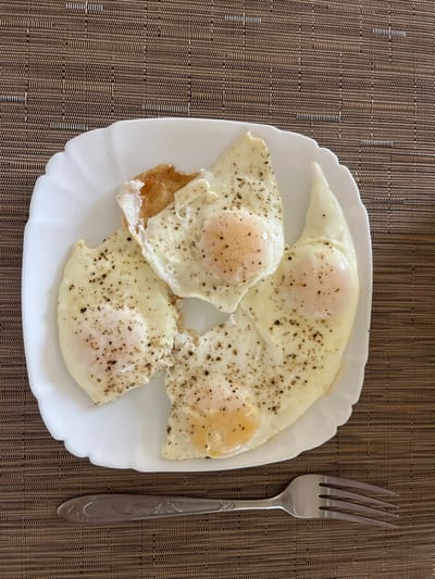 The image presents an overhead view of a culinary scene, stated to be located in the city of Enem, Russia, although no visual elements within the image confirm this geographical setting. A white, square-shaped ceramic plate with scalloped edges dominates the center of the frame, holding four fried eggs. Each egg exhibits a firm, opaque white and a distinct, lightly cooked, pale yellow-orange yolk. All four eggs are uniformly seasoned with coarse black pepper flakes visible across their surfaces. A single silver-colored metal fork, featuring four tines and a decoratively embossed handle, is positioned horizontally beneath the plate, pointing towards the right. The plate and fork rest on a woven mat or surface, characterized by a tight, textured pattern of intertwined brown and beige fibers, creating a subtle grid-like appearance. No individuals, actions, or direct interactions are visible in the scene.