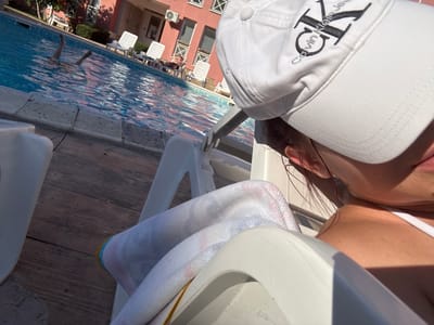 The image captures a sunny day at a poolside in Tankovo, Bulgaria. In the foreground, a person is relaxing on a white lounge chair, partially obscured by a white cap with the Calvin Klein Jeans logo prominently displayed. A white towel with colorful striped edges is draped over the chair.

The background reveals a clear blue swimming pool with several people enjoying themselves. One person's legs are visible kicking in the water, suggesting they are swimming or playing. Further back, more white lounge chairs are set up, with one person lying on a chair, sunbathing. The building behind the pool is a light pink apartment complex with visible windows and an air conditioning unit.

The time of day appears to be midday, with bright sunlight casting shadows and illuminating the scene. The weather is clearly warm and pleasant, ideal for swimming and relaxing by the pool. The overall mood is one of leisure and enjoyment. The visible text on the cap is "Calvin Klein Jeans".