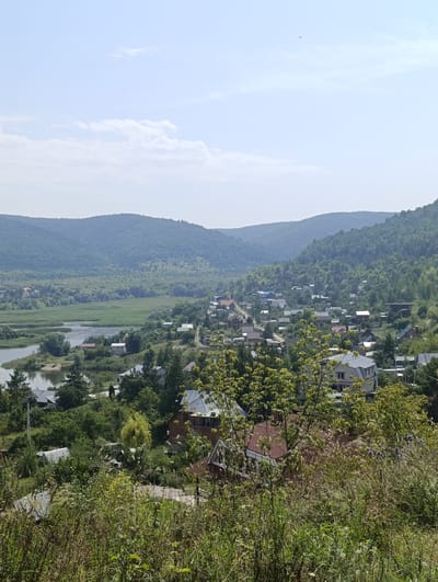 The image shows a panoramic view of the settlement of Solnechnaya Polyana, Russia, on a bright, hazy day. The foreground is dominated by a dense growth of dry grass and green foliage, obscuring the lower part of the scene. Beyond this, a village is nestled among rolling, tree-covered hills. Houses with pitched roofs, varying in color and architectural style, are scattered throughout the landscape. A river or large body of water winds its way through the valley, reflecting the sky and surrounding greenery. In the mid-ground, a lush green field stretches towards the water, bordered by trees. The background consists of several layers of receding, densely forested hills that fade into the hazy horizon. The sky is a pale blue with scattered, wispy white clouds. A small, dark object, possibly an aircraft, is visible high in the sky. The overall impression is of a tranquil, rural settlement set within a verdant natural environment.