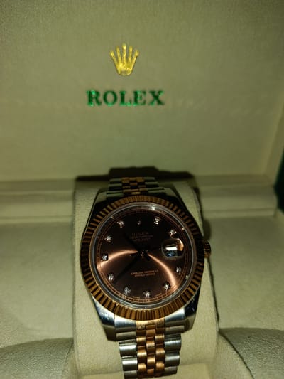 The image shows a close-up of a Rolex watch, presented in a Rolex box. The watch has a two-tone band, combining silver and rose gold. The watch face is a coppery-brown color, adorned with diamond hour markers. The bezel of the watch is fluted and made of rose gold. The date window displays the number "25". The Rolex logo, a gold crown above the word "ROLEX" in green lettering, is embossed on the inside lid of the box. The background of the box is a muted green. The lighting suggests it is indoors, with warm artificial light illuminating the watch and the box. The overall impression is one of luxury and sophistication. There is no indication of the time of day, weather, or specific location beyond the brand of the box and watch. The image is a still photograph, with no activity or event taking place other than the presentation of the watch.