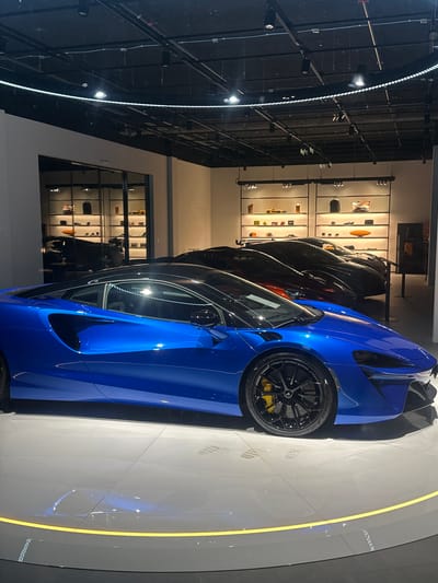 The image shows a car showroom in Glattbrugg, Switzerland. A blue McLaren sports car is prominently displayed on a grey rotating platform outlined with a yellow LED strip. Behind it are several other luxury cars in dark colors. In the background, display cabinets showcase various accessories and merchandise. The showroom has a high ceiling with industrial-style lighting fixtures.