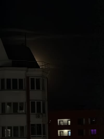 The image depicts a night scene in Bobrovo, Russia, featuring residential buildings under a moonlit sky. In the foreground, a white building with multiple floors and windows is visible. The windows are dark, indicating that the rooms are not lit. An air conditioning unit is mounted on the side of this building. To the right, a red building with a dark facade is also visible, with several windows illuminated by interior lights. One window on the lower right of the red building displays a vibrant purple light. The sky above is dark, with the moon partially obscured by clouds or the silhouette of a building. The upper portion of the white building has a dark, sloped roof and a railing on a balcony or terrace. The scene is devoid of any people or observable activity.