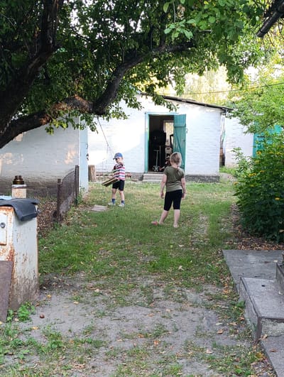 The image depicts a backyard scene in Ozera, Ukraine. Two children are present. A young boy in a striped shirt and shorts stands near a chain-link fence, holding several wooden planks. A young girl, facing away from the camera, wears a green t-shirt and black shorts. They are in a grassy area with a path leading to a white building with teal doors. Various objects, including an old metal structure, a jar, and a scooter, are visible in the background. The environment appears to be a residential backyard with a mature tree overhead, casting shadows.
